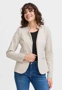 Beige, figurnära blazer med hög krage, slätt tyg och en frampocket, ihop med en svart topp och blå jeans.