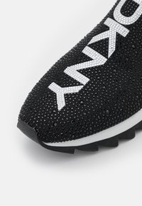 Černá špička tenisky s texturovaným povrchem a bílými kamínkovými nápisy "YMXO" na horní tkanině.