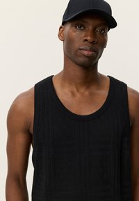 Μαύρο αμάνικο λουράκι με υφασμένα σχέδια, στρογγυλή λαιμόκοψη και φαρδιές μασχάλες, συνδυασμένο με αντίστοιχο μαύρο καπέλο.