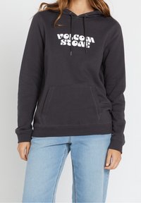 Sweatshirt noir à capuche avec un logo blanc, poche avant et cordon de serrage. Tissu doux avec des poignets et un ourlet côtelés. Associé à un jean en denim clair.