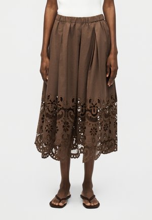 Καφέ μίντι φούστα με λουλουδάτη και παϊλέ μοτίβο δαντελωτό τελείωμα, ελαστική μέση, φορεμένη με λευκό τοπ και καφέ επίπεδα σανδάλια.