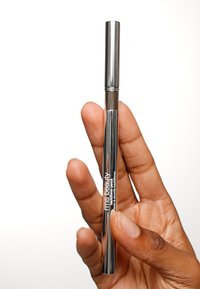 Crayon à sourcils métallique argenté tenu dans une main, avec un design de tube élégant, une étiquette de marque visible et une pointe fine pour précision.
