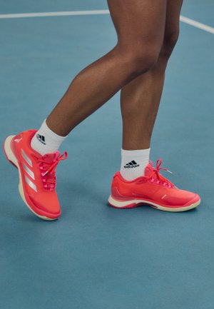 Scarpe da ginnastica di un luminoso colore rosa con accenti e lacci bianchi, caratterizzate da una texture liscia e una suola in gomma, indossate con calze bianche Adidas.