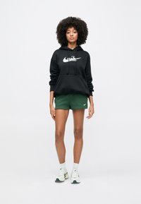 Kvinne stående iført en svart Nike-hettegenser, grønne Nike-shorts, hvite Nike-sokker og hvite joggesko på en ensfarget hvit bakgrunn.