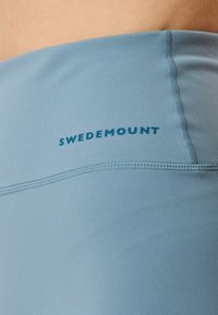 Ljusblå idrottsshorts med en slät yta. Har en sydd söm och "SWEDEMOUNT"-logotypen i mörkblått nära midjan.