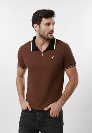 Mann trägt braunes kurzärmeliges Polo mit schwarzem Kragen und Reißverschluss, steht mit den Händen in den Taschen und blickt seitlich vor weißem Hintergrund.