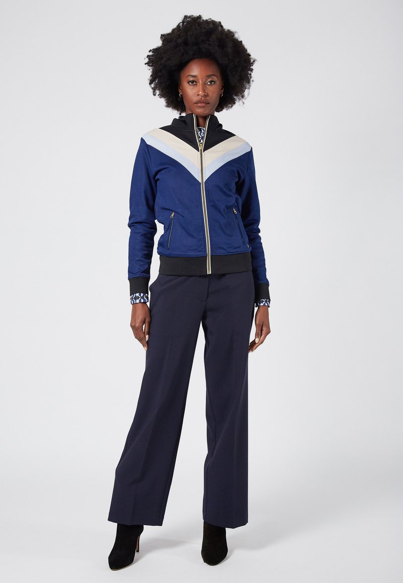 Navy zip-up jacket with a cream and light blue chevron design, featuring side pockets and ribbed cuffs, paired with navy wide-leg trousers.