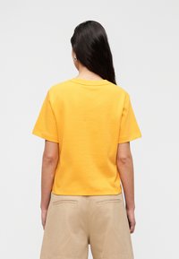 Κίτρινη βαμβακερή μπλούζα με κοντά μανίκια και χαλαρή εφαρμογή. Η πίσω πλευρά έχει λαιμόκοψη τύπου crew και ομαλή υφή. Συνδυάζεται με μπεζ σορτς.