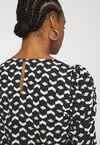 Černý oděv s geometrickým vzorem v bílé a béžové barvě, s nabíranými rukávy, detailem knoflíkové zadní strany a hladkým povrchem.