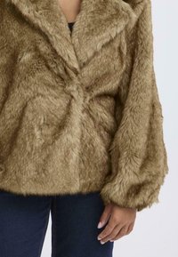 Giacca in pelliccia sintetica marrone con una superficie soffice e texturizzata, maniche ampie e colletto, indossata sopra una maglia blu navy e jeans scuri.