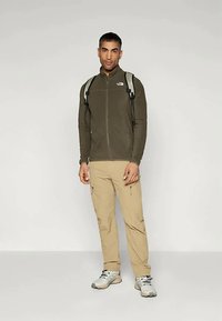 Olive zaļas flīša jaka ar rāvējslēdzēju priekšā; gaiši brūnas vieglas bikses; pelēki pārgājienu apavi; pelēka un melna mugursoma ar siksnām.