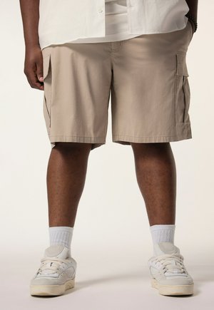 Person med mørk hud, iført beige cargoshorts, hvide sokker, hvide sneakers og en hvid skjorte med den ene hånd i lommen.