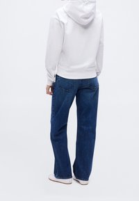 Personne portant un sweat à capuche blanc, un jean droit bleu et des chaussures blanches, debout de dos sur un fond blanc uni.