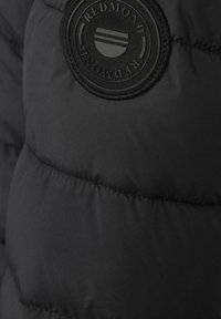 Sort pufferjakke-sektion med et rundt, hævet logo-patch med teksten "REDMOND" og vandrette striber. Glat, mat finish.