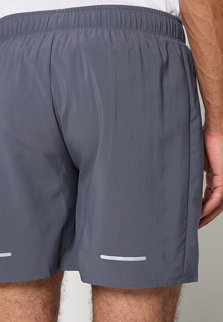 Man in een grijze sportbroek met elastische tailleband en reflecterende strips bij de zoom, staande met zijn arm ontspannen aan de zijkant.