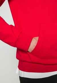 Rotes Sweatshirt mit Kängurutasche, gerippten Bündchen und Abschluss. Über einem weißen Shirt getragen. Glatte Stofftextur.