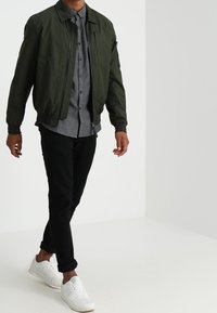 Donkergroene bomberjack, grijs overhemd met knopen, zwarte broek en witte sneakers. Glad stof, ontspannen pasvorm, met opgerolde manchetten en ritszak.