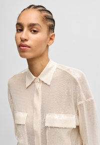 Beige, durchsichtige Button-up-Bluse mit einem strukturierten, glitzernden Finish, die einen kragenlosen Hals und zwei Brusttaschen hat. Legere Passform mit langen Ärmeln.