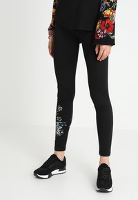 Černé legíny s květinovým vzorem na levé noze, s živými barvami. Doplnené černými sportovními botami. Měkký, elastický materiál.