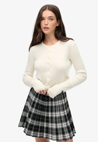 Biały kardigan o splocie w warkocz z długimi rękawami, z okrągłym dekoltem i zapięciem na guziki, w połączeniu z plisowaną spódnicą w czarno-białą kratę.