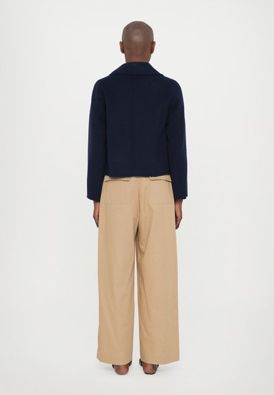 Giacca cropped blu navy con colletto, abbinata a pantaloni larghi beige. Il materiale sembra morbido, con un design minimalista e senza hardware visibile.