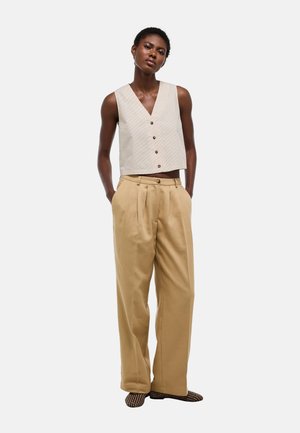 Femme portant un gilet beige sans manches boutonné, un pantalon ample taille haute beige et des chaussures à enfiler à motifs, debout les mains dans les poches.