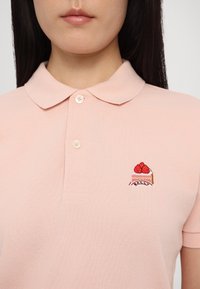 Gros plan d'une personne portant un polo rose pâle avec un petit logo brodé de gâteau à la fraise sur le côté gauche de la poitrine.