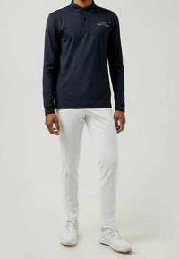 Langärmliges navy Polohemd mit Kragen und einem weißen Logo, kombiniert mit schmal geschnittenen weißen Hosen und weißen Sportschuhen.