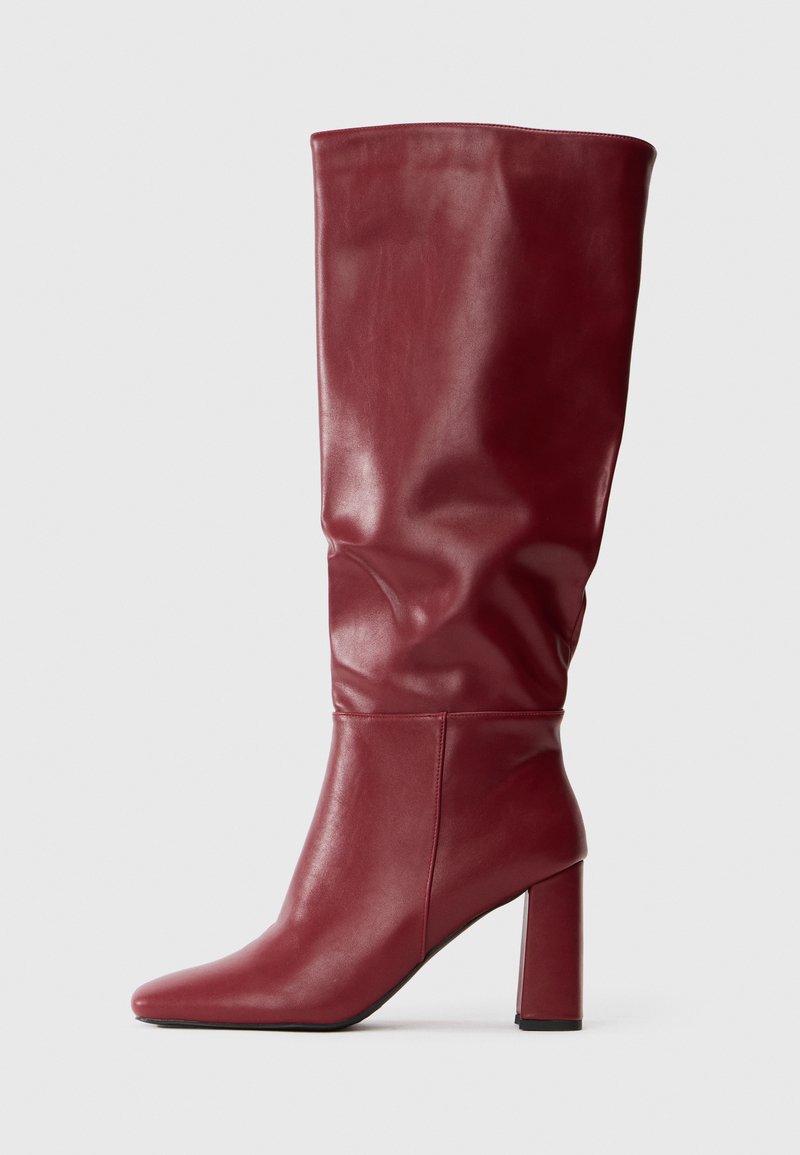 Aukšta bordo odinė batai su laisvu aulu, smailiu pirštu ir aukštu kvadratiniu kulnu, stovintys prieš paprastą šviesų foną.