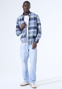 Geklede plaid overhemd in marineblauw, groen en wit; gedragen over een wit T-shirt. Lichtblauwe jeans en witte sneakers maken de look compleet.