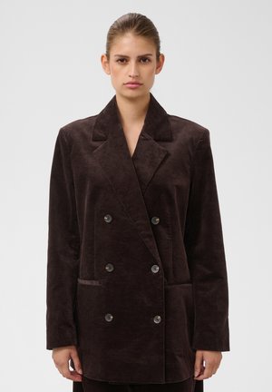 Femme portant un blazer en velours côtelé marron foncé à double boutonnage avec un col cranté, se tenant devant un fond uni clair.