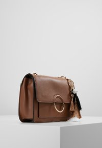 Borsa a tracolla in pelle marrone con patta frontale, hardware dorato, accentuazione ad anello circolare e nappine decorative in bianco e nero.