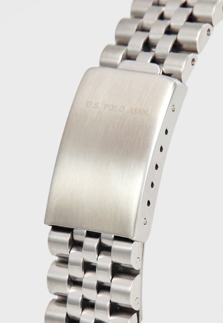 Zilveren roestvrijstalen horloge met een gepolijste rechthoekige wijzerplaat, textuur in de strap schakels en U.S. Polo Assn. gegraveerd op de sluiting.