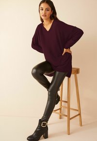 Mujer posando sobre un taburete de madera con una camiseta de manga larga de color púrpura oscuro, leggings de cuero negro y botines negros de tacón con hebillas.