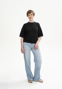 Zwarte gebreide top met korte brede mouwen en horizontale ribbels, gecombineerd met lichtblauwe wijde jeans en grijze sandalen.