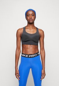 Fekete sportmelltartó struktúrált mintával, amelyhez egy fekete "NIKE PRO" derékpántos, testhez simuló kék leggings illik. A modellen kék hajpánt található.
