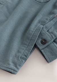 Poignet de manche longue bleu-gris doux avec un bouton noir et une couture latérale visible sur une chemise ou une manche de veste en tissu texturé.