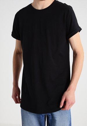 Person iført en ensfarvet sort t-shirt med korte ærmer og opkrøllede manchetter samt lyseblå jeans, stående foran en ensfarvet baggrund.