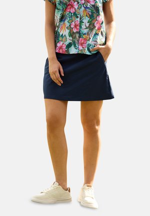 Mujer con una camisa de manga corta con estampado floral, falda azul marino con bolsillos y zapatillas blancas, de pie con una mano en el bolsillo.