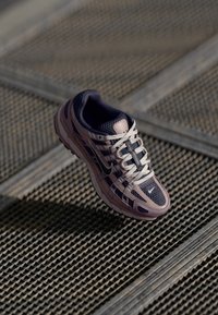 Chaussure de course Nike en mailles violettes et grises, avec des lacets blancs et des superpositions, dotée d'une semelle texturée, sur un fond à motifs.