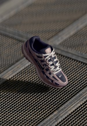 Nike løpesko i lilla og grå mesh, med hvite snører og overlegg, med en teksturert såle, satt mot en mønstret bakgrunn.