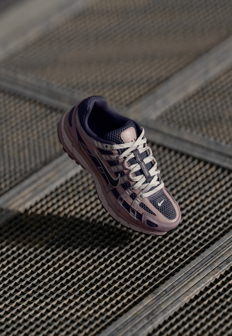 Chaussure de course Nike en mailles violettes et grises, avec des lacets blancs et des superpositions, dotée d'une semelle texturée, sur un fond à motifs.