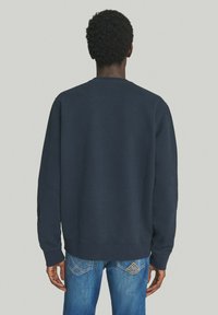 Felpa blu navy con scollatura a girocollo e maniche lunghe, caratterizzata da polsini e orlo a costine. Indossata con jeans azzurri che mostrano dettagli sulla parte posteriore.