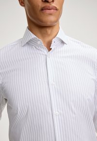 Camicia bianca con sottili righe verticali grigie, con bottoni sul davanti e colletto all'americana. Tessuto di cotone liscio con un design aderente.