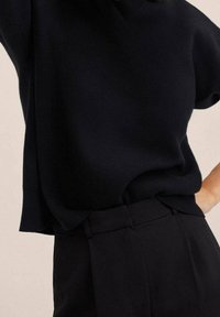 Personne portant un haut noir à manches courtes ample rentré dans un pantalon noir taille haute sur fond beige uni.