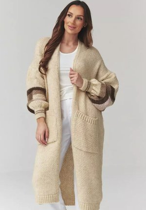 Kobieta w długim beżowym dzianinowym kardiganie z brązowo-kremowymi rękawami w paski typu balloon, białym topie i białych spodniach, pozująca na jednolitym tle.