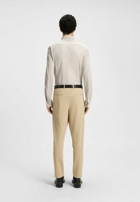 Lichtbeige langemouwshirt met een gladde textuur, gecombineerd met textuurbeige broeken en zwarte schoenen, zichtbaar vanuit de achterkant.
