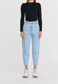 Frau trägt ein schwarzes langärmliges Oberteil mit Puffärmel, hellblaue hochgeschnittene tapered Jeans und weiße klobige Sneaker.