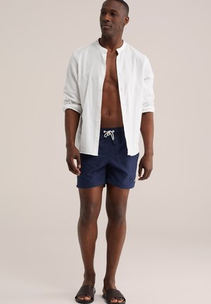 Muž v bielej rozopnutej košeli na gombíky, námorníckych šortkách so šnúrkou a tmavých tkaných šľapkách, stojaci pred neutrálnym pozadím.