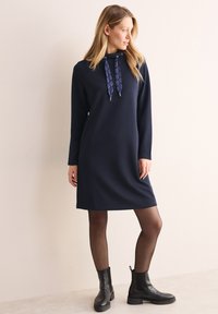Abito a righe blu navy con cappuccio, dotato di cordini decorati. Il modello indossa stivali neri alla caviglia e collant neri trasparenti.